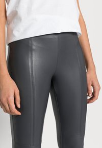 Hoge taille donkergrijze nepleer legging met een gladde textuur en zichtbare stiknaaddetails, gecombineerd met een wit katoen T-shirt.
