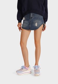 Sneakers grigio chiaro e pastello con suola chunky, caratterizzati da un design curvo, sovrapposizioni testurizzate e lacci coordinati. Indossati con una minigonna di denim.