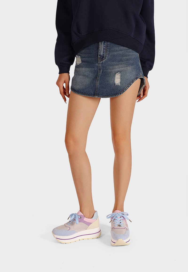 Sneakers grigio chiaro e pastello con suola chunky, caratterizzati da un design curvo, sovrapposizioni testurizzate e lacci coordinati. Indossati con una minigonna di denim.