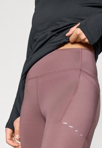 Leggings mauves à texture lisse, avec des coutures plates et des bandes d'accent blanches. Portés avec un haut noir à manches longues.