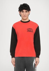 Crvena i crna majica s dugim rukavima, okruglog izreza i malim grafičkim logotipom na prednjoj strani. Materijal mješavine pamuka s opuštenim krojem.