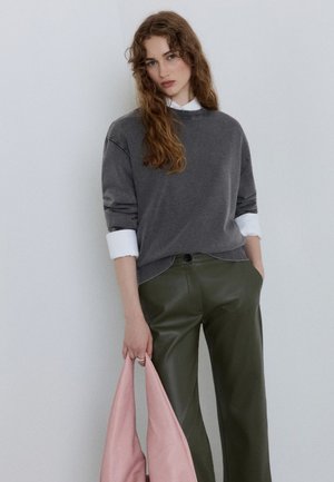 Giovane donna con capelli lunghi e ricci che indossa un maglione grigio sopra una camicia bianca e pantaloni verdi, tenendo una borsa rosa contro un muro uniforme.