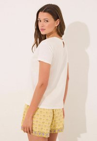 Witte top met korte mouwen en een klein keyhole-detail aan de achterkant, gecombineerd met gele shorts met bloemenpatroon en blauwe accenten. Zachte katoenen stof.