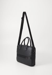 Black leather bag with a textured surface, dual top handles, and a long adjustable strap. Features a sleek design and matte accents.