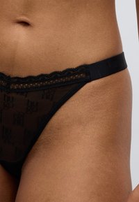 Μαύρο δαντελένιο στρινγκ με κυματιστά άκρα και ελαστική μέση. Διαθέτει διαφανή υφή και μικρή λεπτομέρεια με λογότυπο.