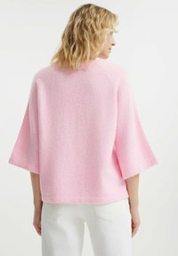 Maglione rosa fluffy con maniche ampie e accorciate e un collo alto. Texture morbida, orlo piatto e vestibilità oversize. Indossato con pantaloni bianchi.