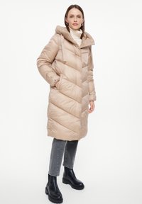 Beige pufferjas met een capuchon, voorzien van een chevronpatroon, zijzakken en een glanzende textuur. Gekozen in combinatie met grijze jeans en zwarte chunky laarzen.