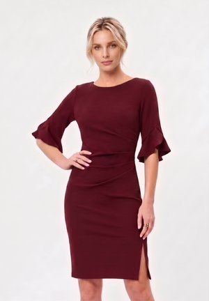 Femme aux cheveux blonds portant une robe bordeaux ajustée avec des manches à volants jusqu'aux coudes et une fente latérale, posant avec une main sur la hanche.
