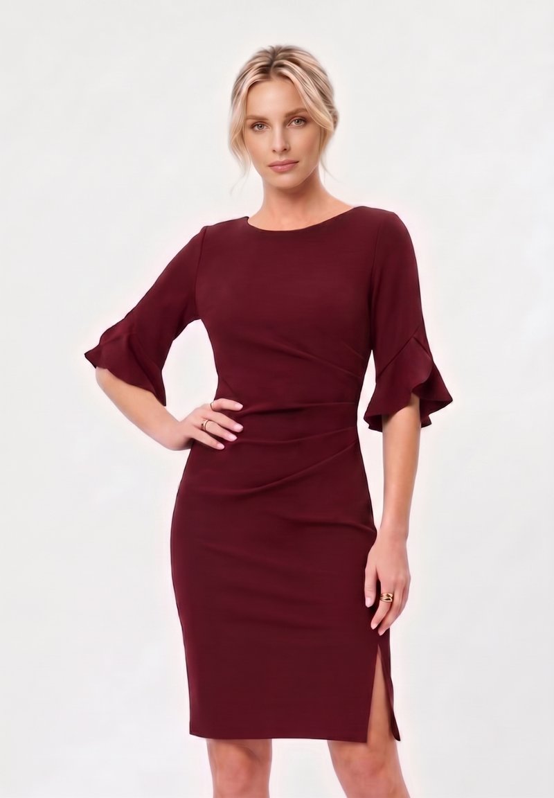 Femme aux cheveux blonds portant une robe bordeaux ajustée avec des manches à volants jusqu'aux coudes et une fente latérale, posant avec une main sur la hanche.