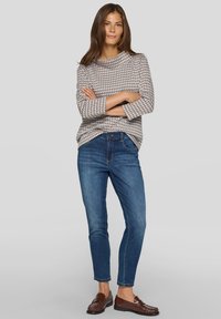Frau mit langen braunen Haaren, die ein gemustertes beige und weißes Oberteil, blaue Jeans und braune Mokassins trägt, mit verschränkten Armen dasteht.