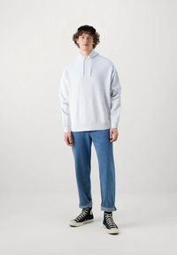 Világosszürke kapucnis pulóver magas nyakkal és állítható húzózsinórral, kék farmernadrággal és fekete magas szárú tornacipővel párosítva.