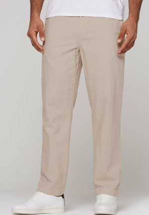 Mann trägt beige Leinenhosen mit weißen Sneakers und einem weißen Hemd, steht vor einem einfarbigen Hintergrund.