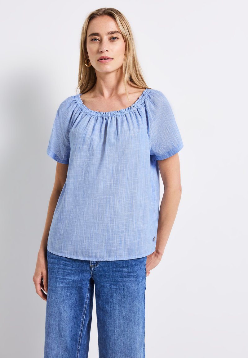 Top azzurro chiaro a maniche corte con scollo arricciato, realizzato in un tessuto testurizzato. Abbinato a jeans blu a gamba larga, che mostrano una vestibilità rilassata.