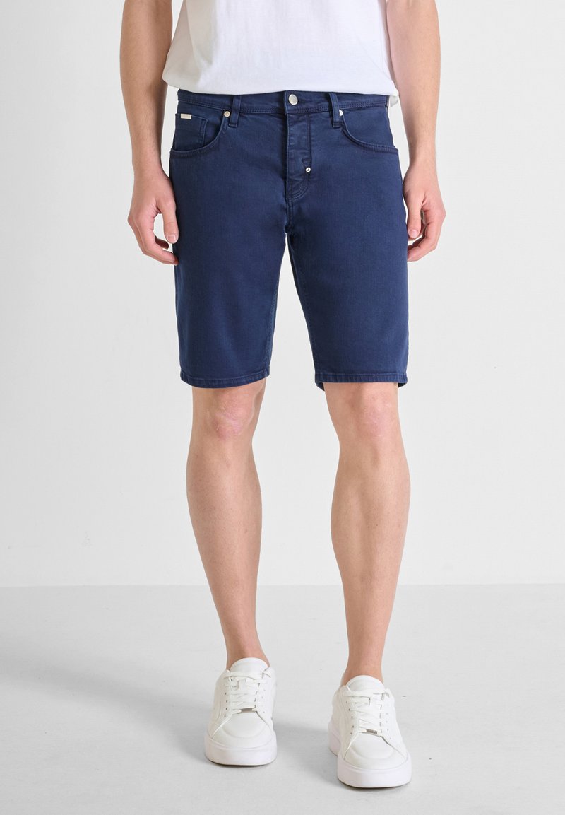 Shorts de mezclilla azul marino con cinco bolsillos, trabillas para el cinturón y cierre de botón. Combinados con una camiseta blanca simple y zapatillas blancas.