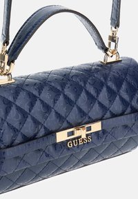 Sac à main matelassé bleu marine avec une texture brillante, doté d'un fermoir doré et d'un marquage. Comprend deux poignées courtes pour le transport.