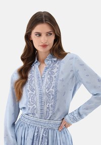 Light blue blouse with a patterned placket and sleeves, features paisley motifs and embroidery, soft fabric, tailored fit, and a button front.