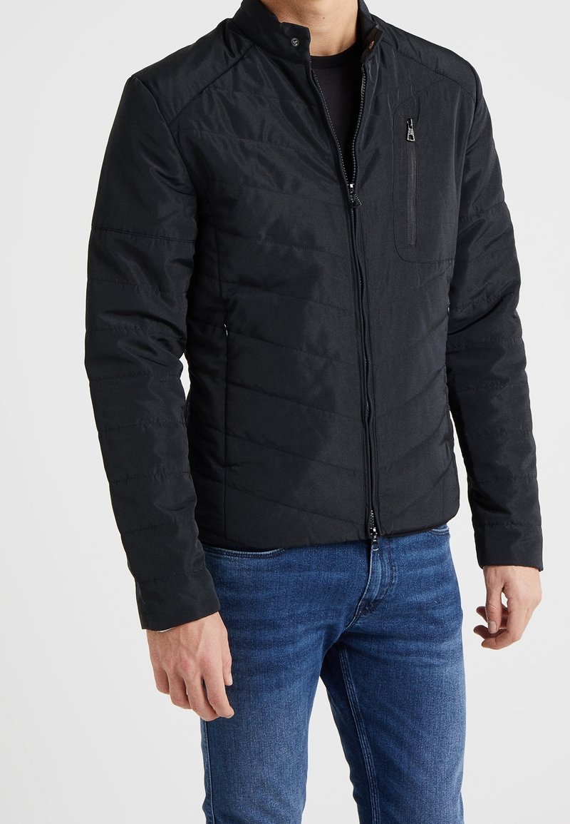 Czarna pikowana kurtka z kołnierzem stójkowym, zapięciem na zamek błyskawiczny oraz przednią kieszenią. Posiada gładką, matową fakturę i dopasowany krój. Połączona z niebieskimi dżinsami.