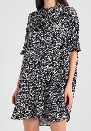 Frau trägt ein locker sitzendes schwarzes und weißes Kleid mit kurzem Ärmel, Mandarin-Kragen und durchgehender Knopfleiste vorne.