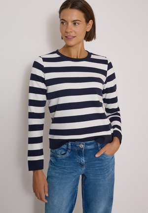 Femme portant un t-shirt à manches longues à rayures horizontales bleu marine et blanc et un jean bleu, debout avec une main dans la poche devant un fond uni.