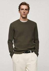 Olivgrüner Rippstrickpullover mit Rundhalsausschnitt, langen Ärmeln und eng anliegendem Saum, getragen über einem weißen Hemd, kombiniert mit beigen Hosen.