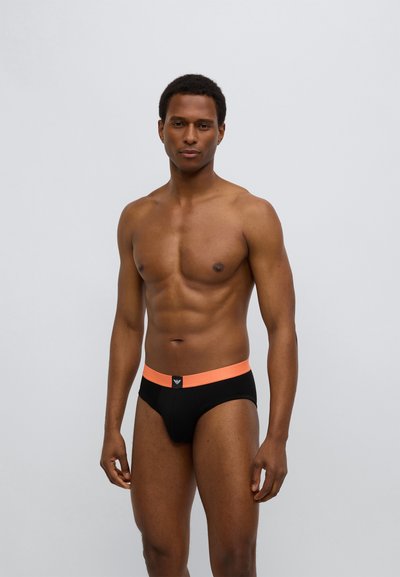 Schwarze Slips mit einem orangen Bund, der ein Logo zeigt. Das Material wirkt glatt mit einem figurbetonten Schnitt. Das Modell steht in einer neutralen Pose.