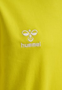 Ljust gul t-shirt i mjukt tyg med en broderad vit Hummel-logotyp och ett distinkt mönster på bröstet.