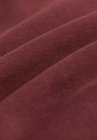 Burgundia soonik kangas, millel on sile tekstuur, esitleb horisontaalseid jooni ja pehmeid voldikesi, mis viitavad venivusele ja paindlikkusele.