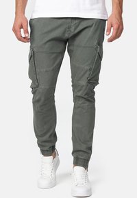 Homme portant un pantalon cargo gris avec plusieurs poches et des poignets élastiques, associé à des baskets blanches sur un fond uni.