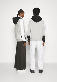 Due persone stanno schiena contro schiena, indossando una felpa grigia con cappuccio e dettagli in bianco e nero e pantaloni neri, in contrasto con una lunga gonna nera.
