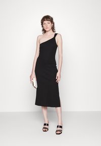 Vestido preto justo, com uma alça única, de comprimento intermédio, apresentando uma fenda lateral, combinado com uma clutch prateada texturizada e sandálias pretas.