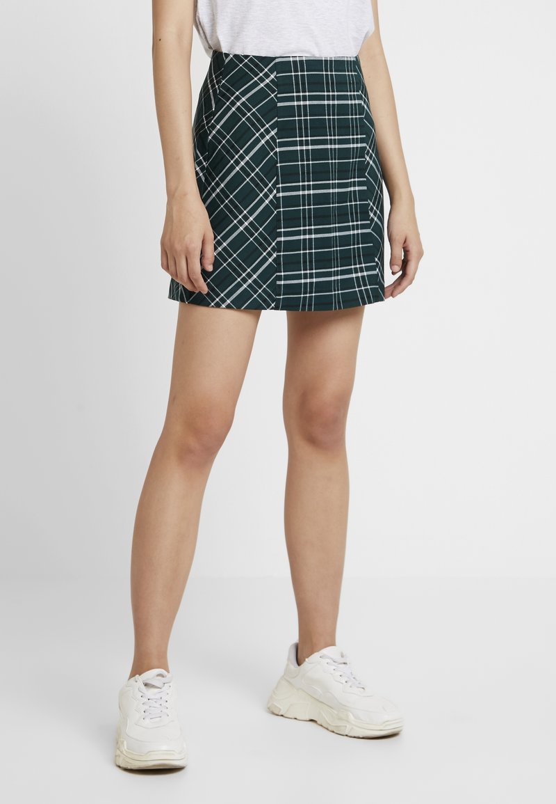 Mini-jupe écossaise verte à fines lignes blanches, avec une taille ajustée et une coupe droite. Portée avec un t-shirt gris décontracté et des baskets blanches.