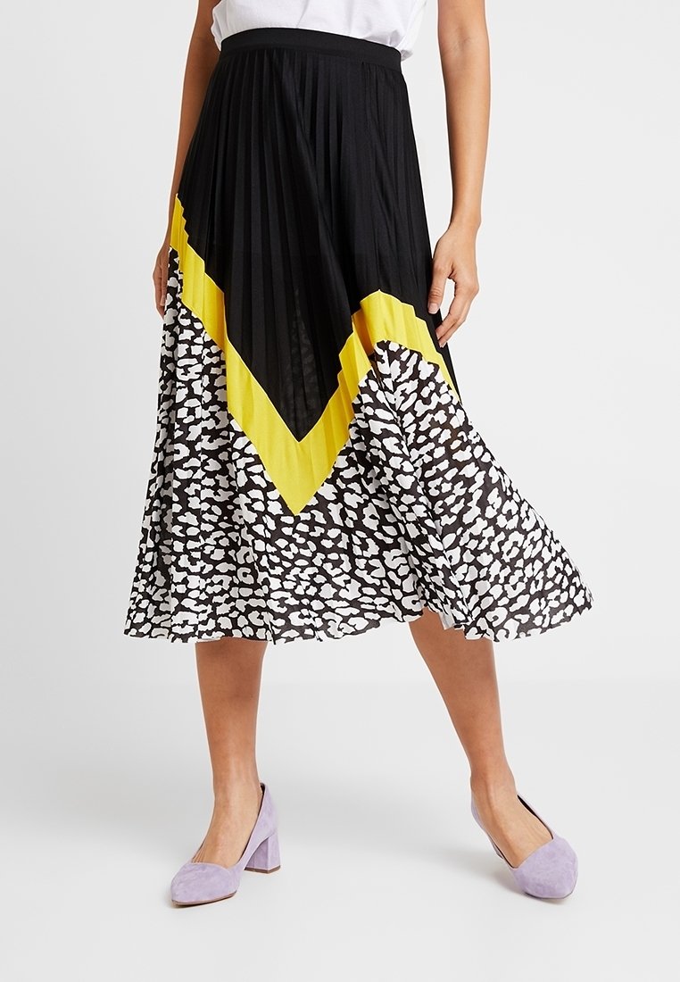 Gonna nera plissettata con motivo a chevron giallo in vita e metà inferiore con stampa animalier bianca. Tessuto leggero. Indossata con scarpe lavanda.
