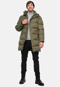 Olivengrøn pufferjakke med lynlås foran, justerbar hætte og ribbede manchetter. Båret over en grå turtleneck, parret med sorte jeans og støvler.