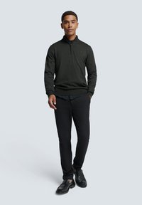 Donkergroen gebreid vest met een ritssluiting bij de kraag, gecombineerd met zwarte broek en zwarte leren schoenen. Eenvoudig ontwerp, zachte textuur en aansluitende pasvorm.