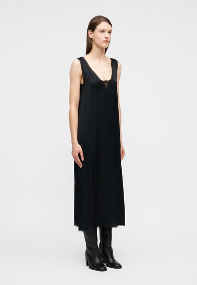 Γυναίκα που φοράει μαύρο midi φόρεμα χωρίς μανίκια με δαντελωτή διακόσμηση και μαύρες μπότες με τακούνι, στέκεται μπροστά σε απλό ανοιχτόχρωμο φόντο.