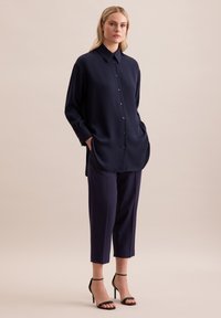 Camicia blu navy oversize con colletto, dotata di due tasche laterali; abbinata a pantaloni blu navy a sigaretta e sandali neri con tacco.