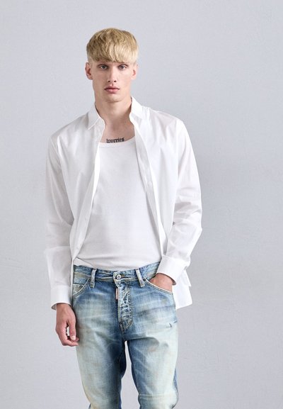 Persona bionda con i capelli corti che indossa una camicia bianca aperta sopra una canotta bianca e jeans azzurri sbiaditi, in piedi contro un muro chiaro e uniforme.