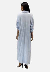 Blue, long-sleeved dress made of textured fabric, featuring a relaxed fit, collar, and rolled sleeves, extending to the ankles. Worn with black heels.