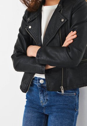 Svart konstläderbikerjacka med dragkedjefickor och slagkrage. Buren över en vit skjorta och blå denimjeans. Detaljerad sömnad synlig.