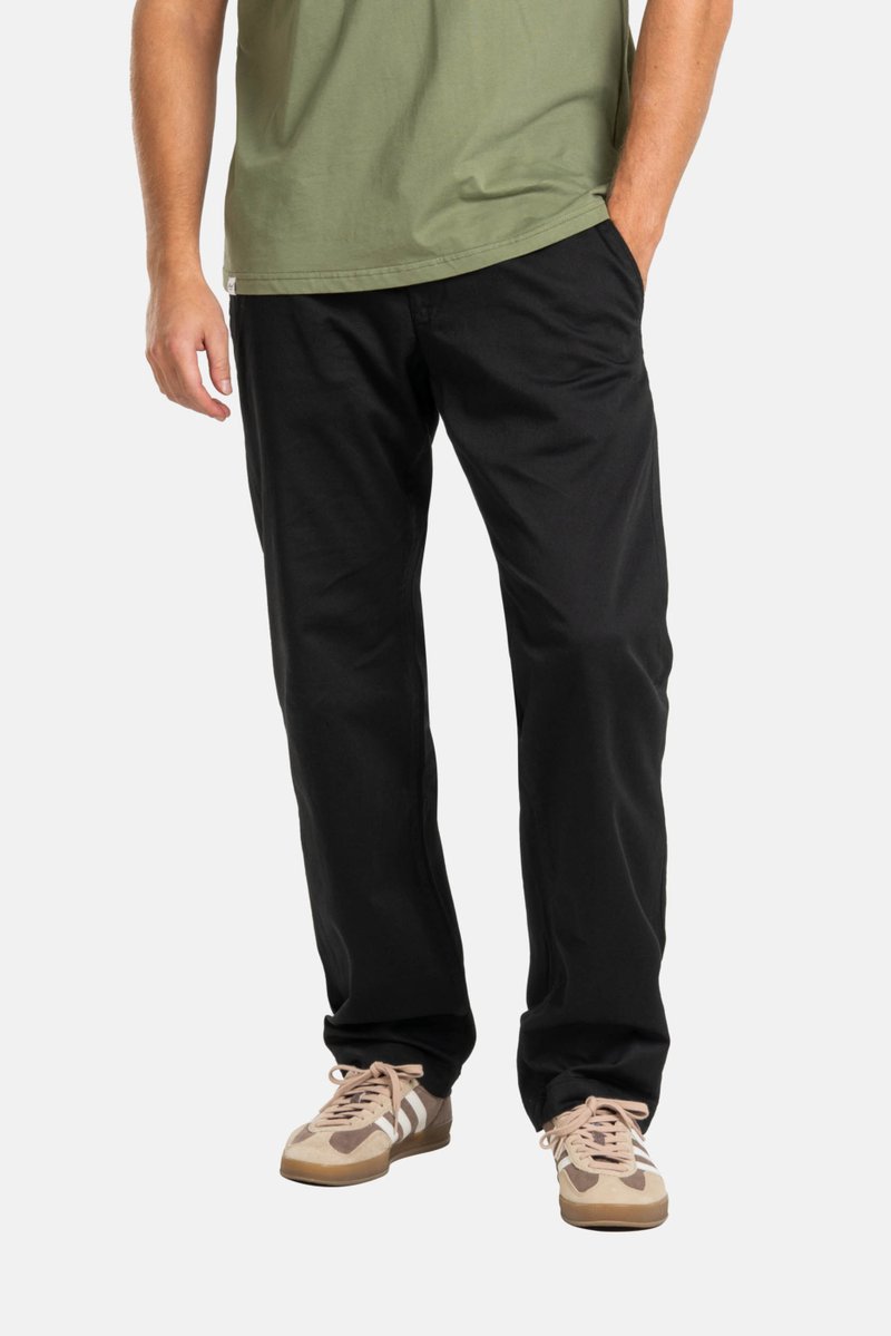 Pantalones de algodón negros con un corte relajado, frontal plano y bolsillos laterales, combinados con una camiseta verde y zapatillas deportivas beige.