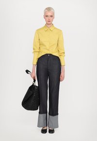 Chemise jaune à manches longues, assortie à un pantalon en denim noir à jambes larges avec des ourlets gris. Sac à main en osier noir et ballerines noires.