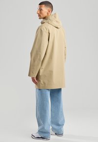 Beige waterproof coat with a hood, long sleeves, and a relaxed fit. Paired with loose-fit light blue denim jeans and white sneakers.