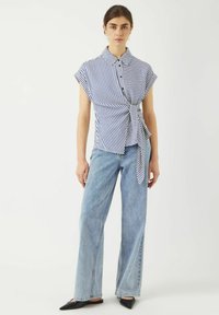 Blusa a righe con maniche corte, chiusura a bottoni e dettaglio a fiocco, abbinata a jeans a vita alta blu chiaro con gamba larga e scarpe nere a punta.