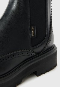 Black leather ankle boot with elastic side panels, brogue detailing along the toe, and a chunky black sole. Features a gold label.