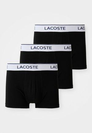 Svarta bomullsboxer med vit elastisk midjeband som har "LACOSTE"-logotyp. Tre par prydligt staplade. Slät textur, klassisk design.