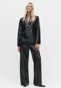 Nainen, jolla on pitkät vaaleat hiukset, pukeutuneena mustaan tekonahkableiseriin ja leveälahkeisiin housuihin, seisoo yksinkertaista vaaleaa taustaa vasten.