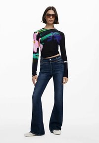 Crna majica dugih rukava s šarenim cvjetnim uzorkom na lijevoj ruci, uparena s tamnoplavim trapericama proširenog kroja i bijelim cipelama. Glatka tekstura, moderan kroj.