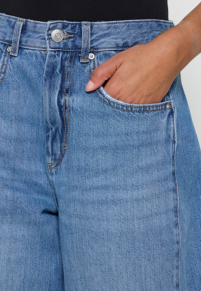 Jasnoniebieskie dżinsy z wysokim stanem, w stylu pięciu kieszeni, z subtelną, zblakłą teksturą. Widoczne detale metalowego guzika i przeszycia.