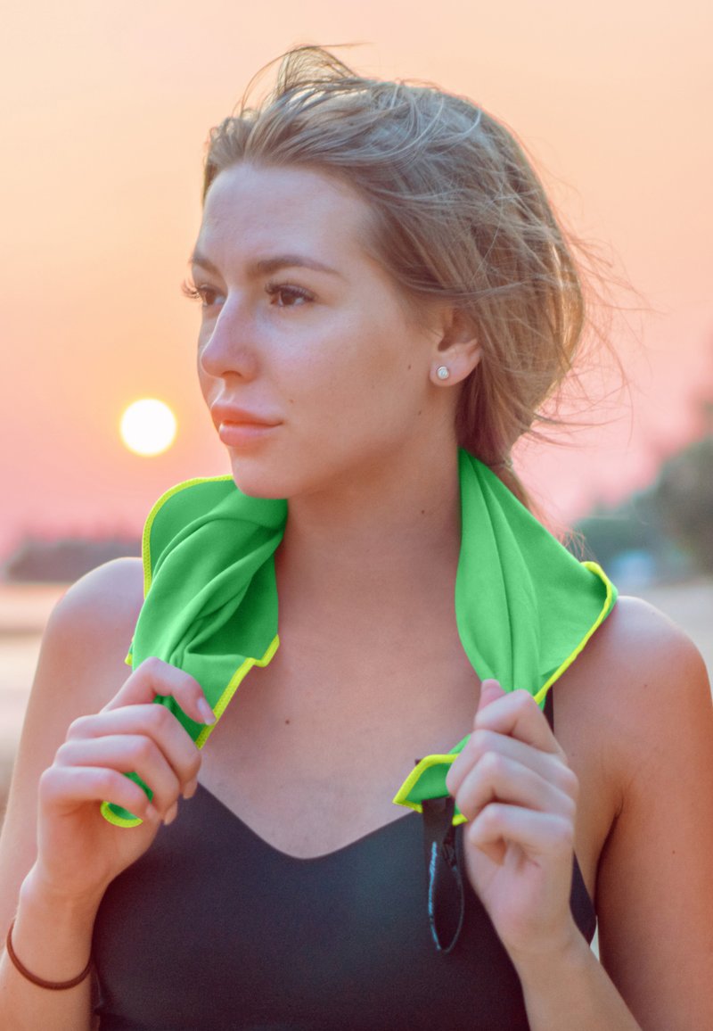 Bright green towel with yellow trim, draped around the neck, worn over a black swimsuit. The background features a sunset and blurred shoreline.