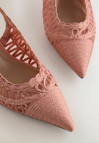 Roze platte schoenen met spitse neus, geweven materiaal, gevlochten details en open zijkanten, op een witte achtergrond.